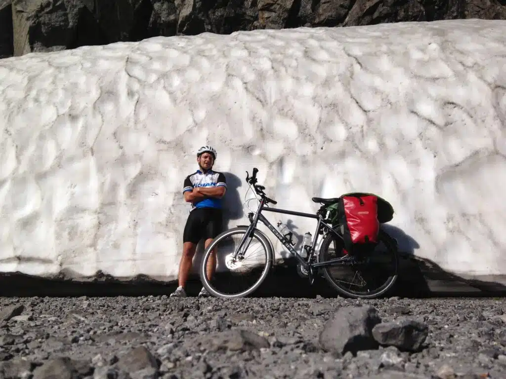 Paul with his loaded touring bike next to a wall of snow, July 2013