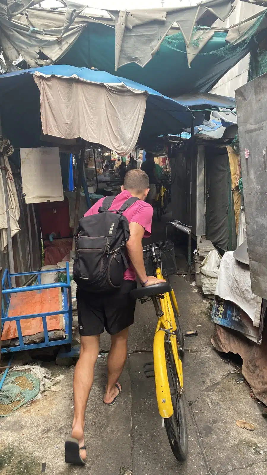 Pushing a yellow Co van Kessel bike through a narrow covered alley with small shops in Bangkok
